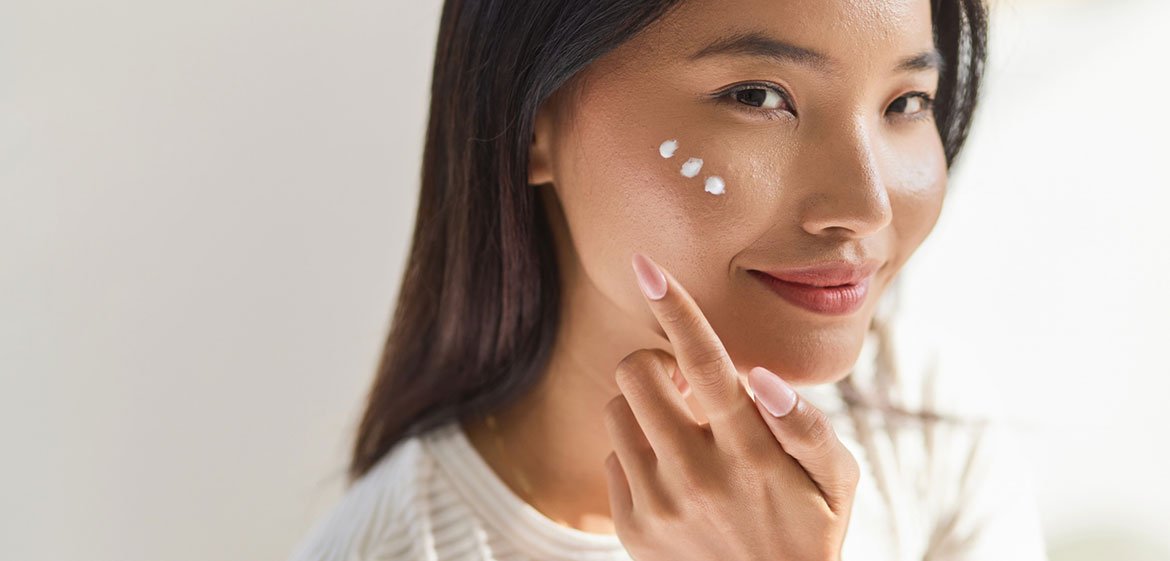 Lächelnde Frau schaut in die Kamera und zeigt auf drei Punkte Creme auf ihrer Wange.